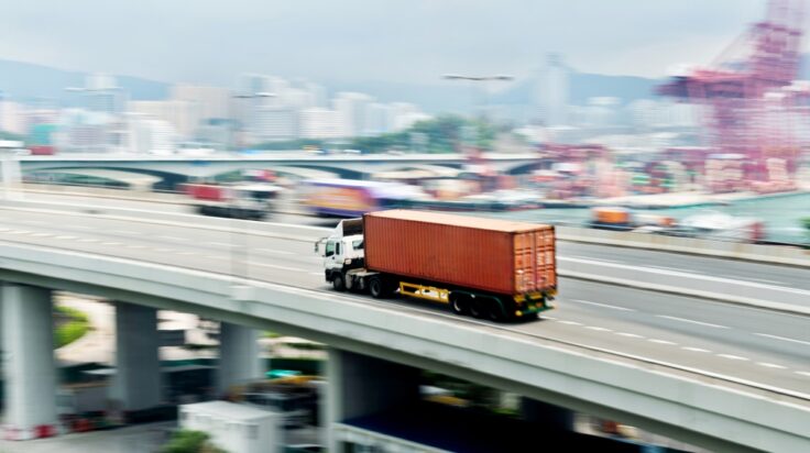 Truck carrying a red shipping container drives on an elevated highway with a distant city skyline and port cranes behind it.