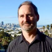 Man outdoors with a city skyline and palm trees in the background; gray-haired, bearded man wearing a dark shirt looking at the camera.