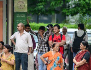 Group of Indian people waiting at a bus stop, with a green bus routes sign and clock behind them.