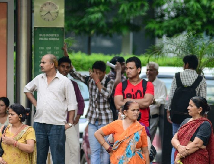 Group of Indian people waiting at a bus stop, with a green bus routes sign and clock behind them.