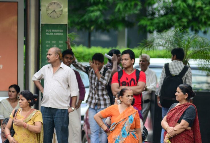 Group of Indian people waiting at a bus stop, with a green bus routes sign and clock behind them.
