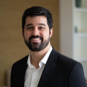 Portrait of a smiling man in a black blazer and white shirt, indoors with a neutral background.