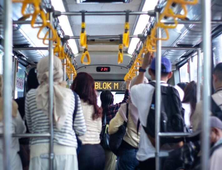 Crowded bus interior with standing passengers gripping yellow handrails; LED display reads 'BLOK M' at the front.