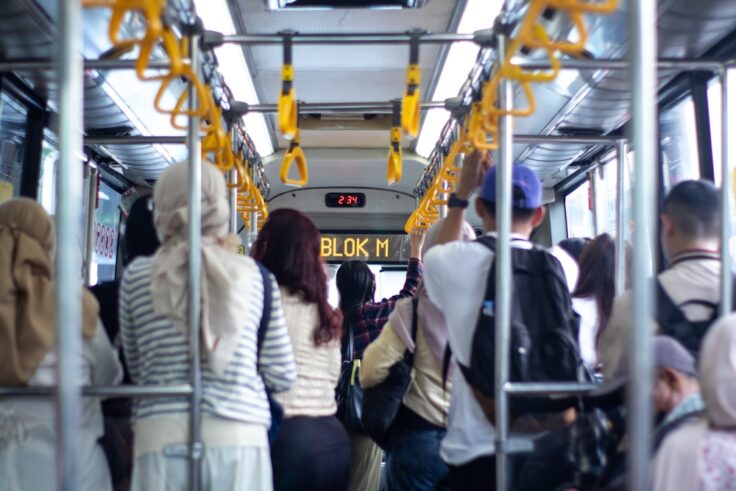 Crowded bus interior with standing passengers gripping yellow handrails; LED display reads 'BLOK M' at the front.