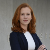 Professional woman with red hair wearing a navy blazer and white blouse, standing with arms crossed against a concrete background