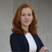 Professional woman with red hair wearing a navy blazer and white blouse, standing with arms crossed against a concrete background