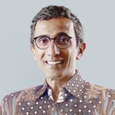 Smiling man with round glasses and short salt‑and‑pepper hair in a patterned shirt, against a gray backdrop.