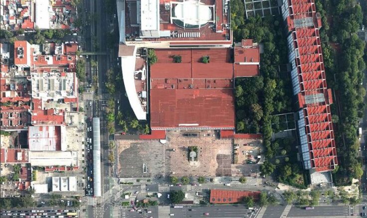 Aerial view of a dense urban block with red roofs, a central open area, surrounding streets, and green trees along the edge.