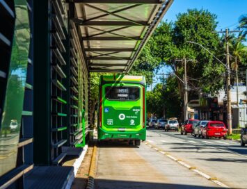Green city bus stopped at a curb beside a modern glass-fronted building on a sunny street.