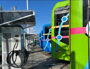 Colorful electric buses lined up at an outdoor charging depot with cables plugged into chargers on a sunny day.