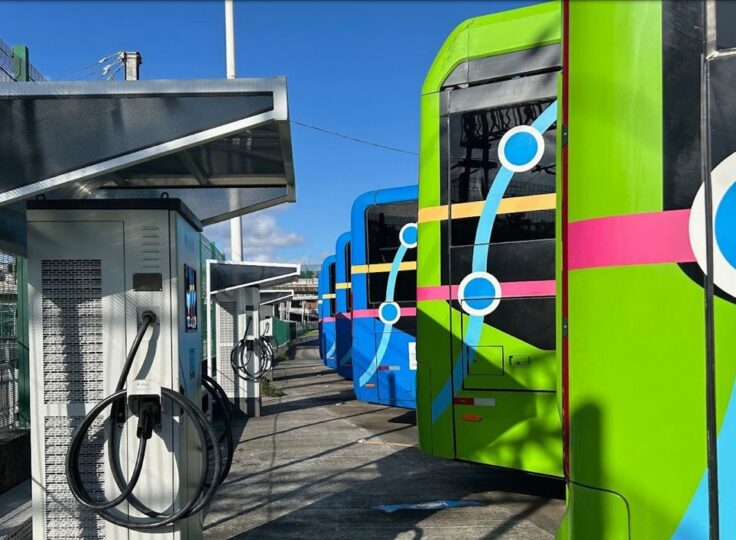 Colorful electric buses lined up at an outdoor charging depot with cables plugged into chargers on a sunny day.