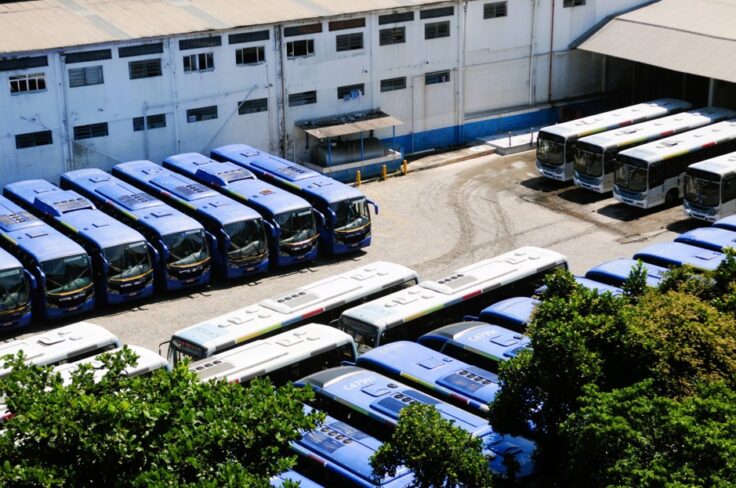 Fleet of blue and white buses parked in neat rows at a bus depot beside a large industrial building and trees in the foreground.