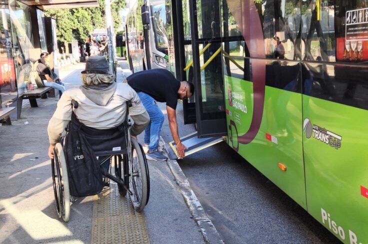 Wheelchair user at a bus stop with a helper preparing to board a green transit bus adjacent to the curbside platform.
