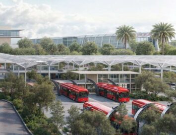 Red buses parked at a modern transit hub with glass canopies and palm trees on a sunny day.