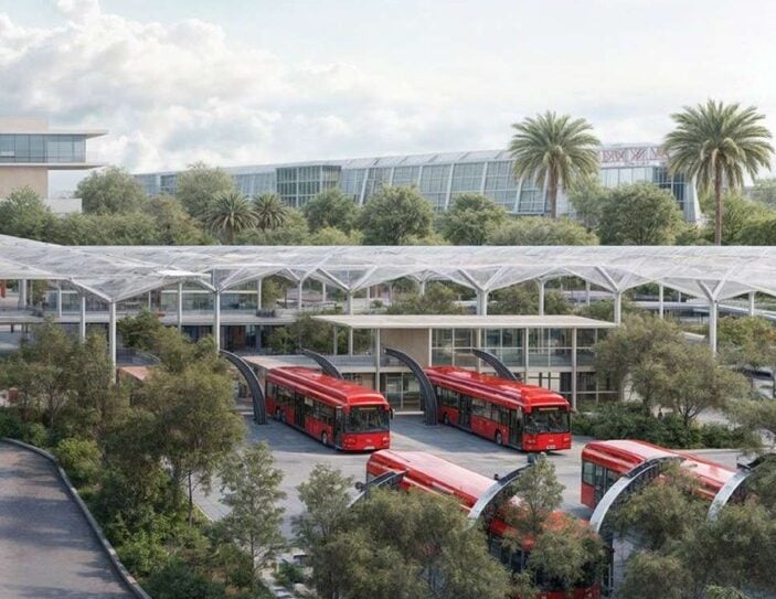 Red buses parked at a modern transit hub with glass canopies and palm trees on a sunny day.