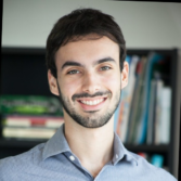 Smiling man with dark hair and beard, wearing a light blue button-down shirt, in front of a bookshelf.