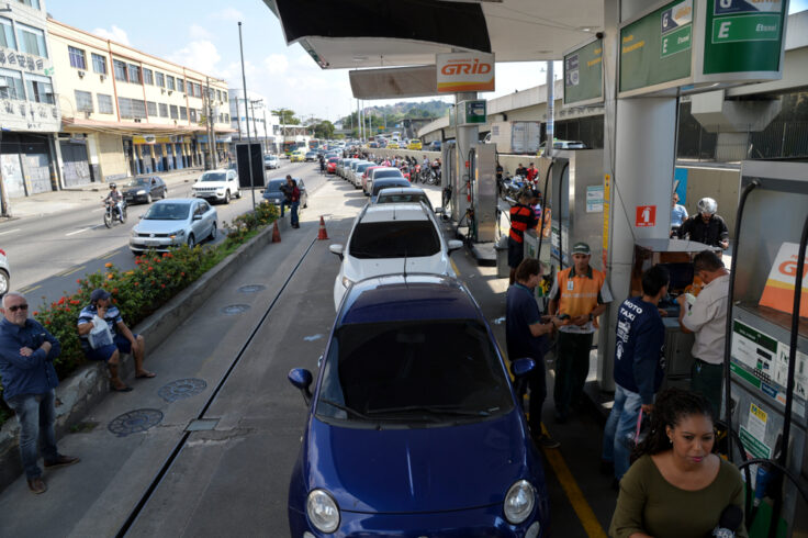 Busy gas station with a long queue of cars refueling under a canopy; a blue car is at the camera in the foreground as attendants assist customers.