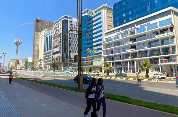 Two young women walk along a wide sidewalk in front of modern glass office buildings and light traffic on a city street.