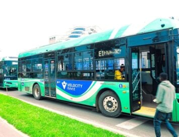 Green city bus with Velocity branding parked at the curb as a passenger boards through the open door.