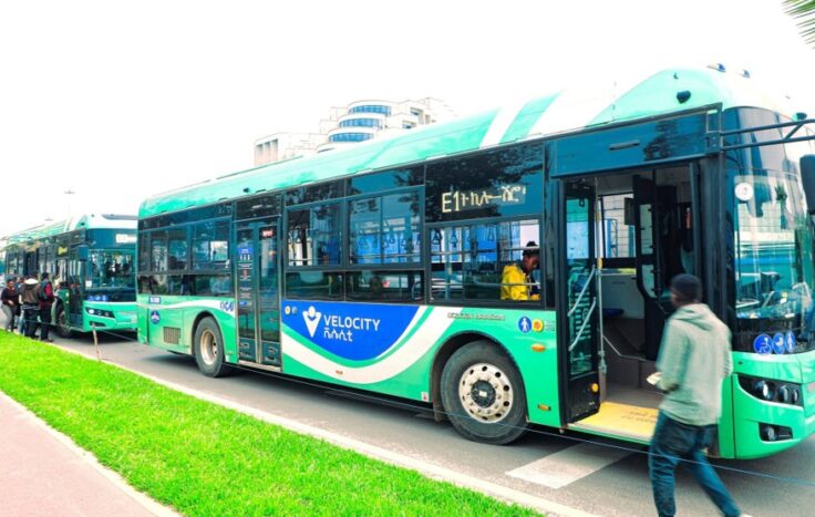 Green city bus with Velocity branding parked at the curb as a passenger boards through the open door.