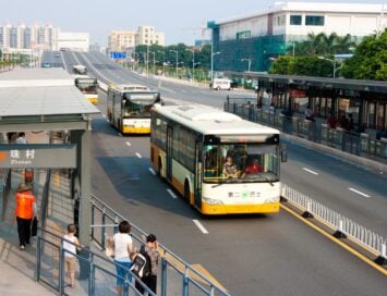 Bus stop scene in Zhucun with waiting passengers as yellow-and-white buses travel on a wide multi-lane road with an urban skyline in the background.
