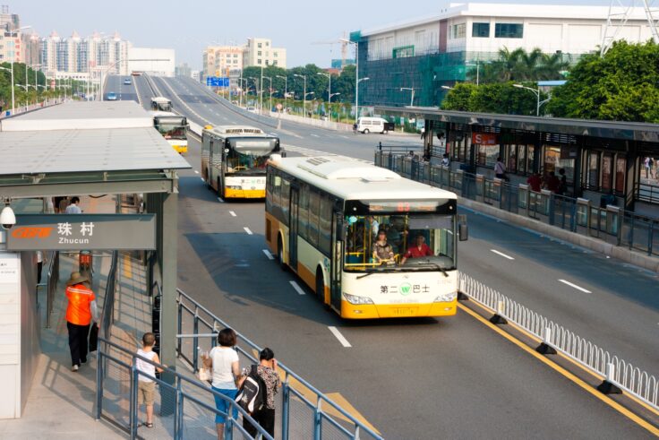 Bus stop scene in Zhucun with waiting passengers as yellow-and-white buses travel on a wide multi-lane road with an urban skyline in the background.