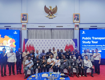 Group of professionals posing for a group photo at a Public Transport Study Tour event, with blue screens and flags in the background.