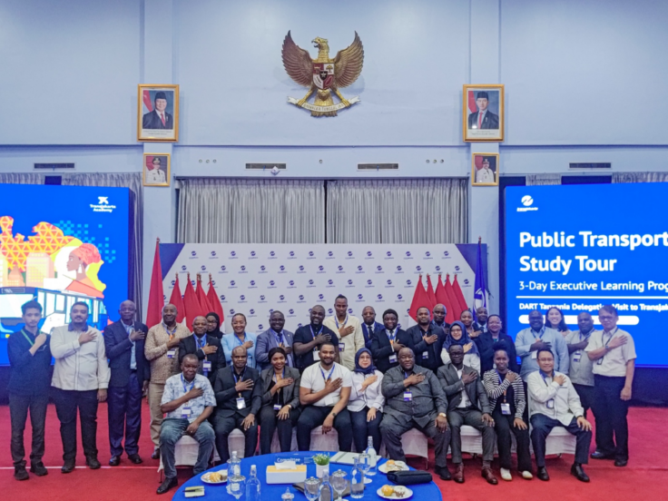 Group of professionals posing for a group photo at a Public Transport Study Tour event, with blue screens and flags in the background.