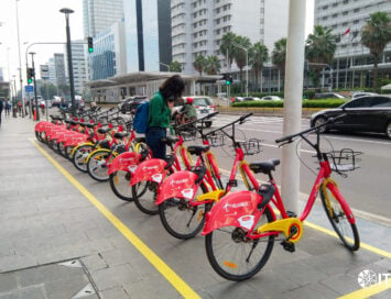 Row of red Telkomsel rental bicycles lined up along a city sidewalk as a woman in a green shirt checks her phone nearby a busy street corner