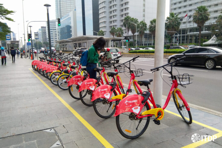 Row of red Telkomsel rental bicycles lined up along a city sidewalk as a woman in a green shirt checks her phone nearby a busy street corner