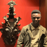 Young man posing beside a large ornate wooden mask sculpture mounted on a red gallery wall.