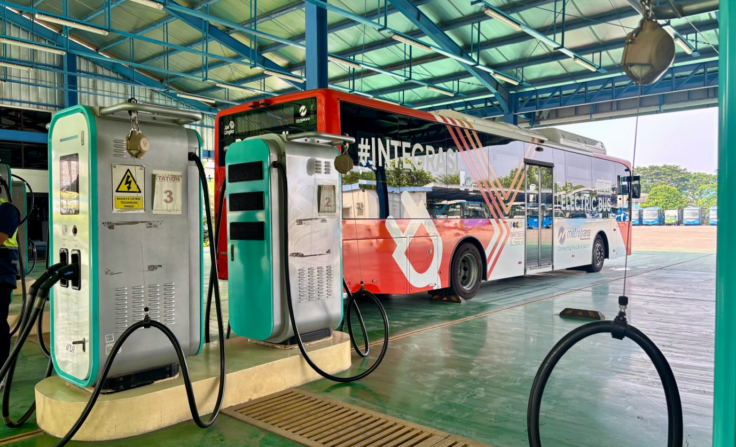 Electric bus connected to a teal charging unit at a depot, with blue structural beams and other buses visible in the background.
