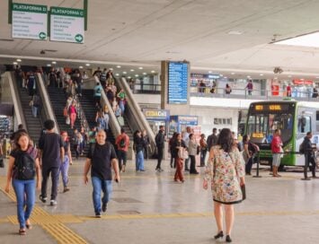 Crowded bus/rail terminal interior with escalators in the center, overhead signs for Plataforma D and C, and a green city bus on the right as passengers move through the concourse.