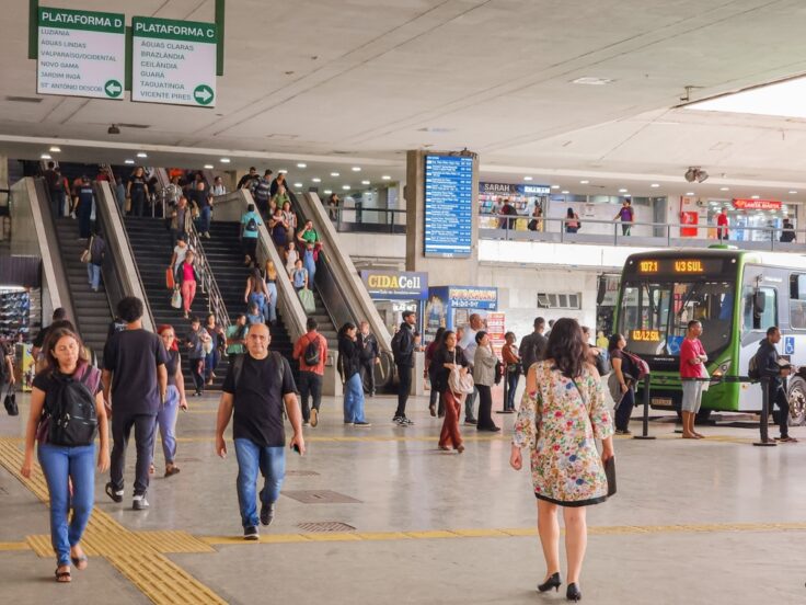 Crowded bus/rail terminal interior with escalators in the center, overhead signs for Plataforma D and C, and a green city bus on the right as passengers move through the concourse.