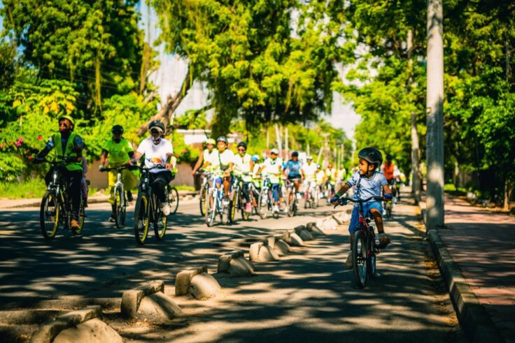 A line of cyclists rides down a tree-lined street, with a smiling child on a bike in the foreground on the right.