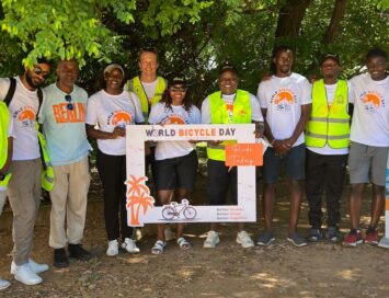 Group of people in safety vests and white World Bicycle Day shirts posing with a decorative frame banner.