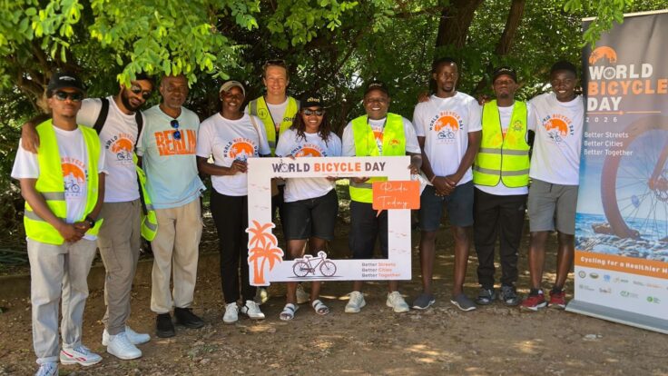 Group of people in safety vests and white World Bicycle Day shirts posing with a decorative frame banner.