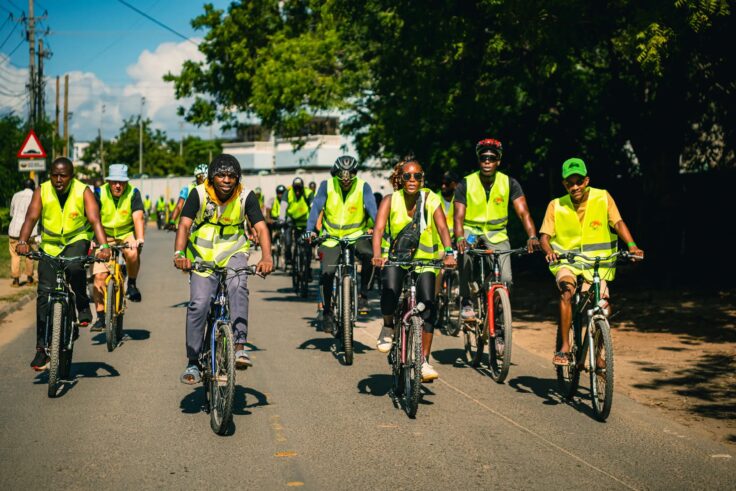 Group of cyclists in high-visibility vests riding together on a sunlit urban street.