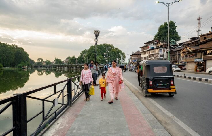 Family strolling along a riverside walkway with a canal on the left and a busy street on the right; a woman in pink leads two children.