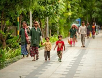Group of women and children walking along a tree-lined sidewalk; two small children hold hands with adult women wearing green tunics and patterned skirts.