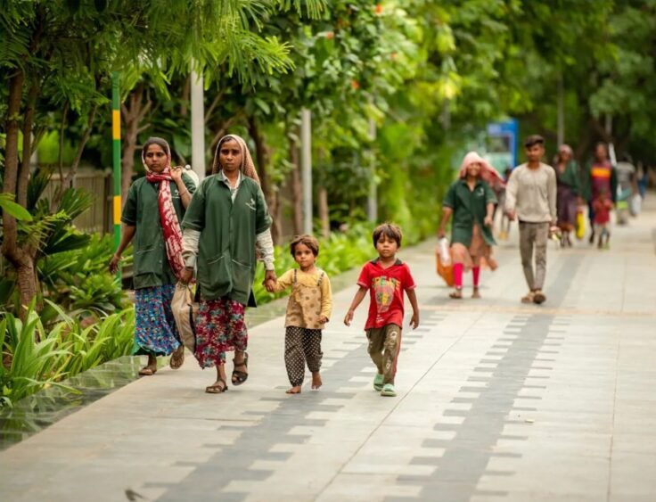Group of women and children walking along a tree-lined sidewalk; two small children hold hands with adult women wearing green tunics and patterned skirts.