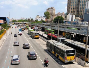 Aerial view of a busy city street with many buses and cars, a bus terminal on the right, and tall buildings in the background.