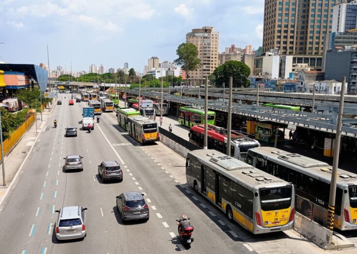 Aerial view of a busy city street with many buses and cars, a bus terminal on the right, and tall buildings in the background.