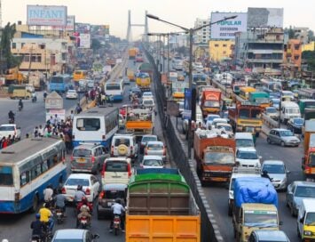 Congested urban highway at rush hour with cars, buses, and trucks crowded in multiple lanes while pedestrians stand on the roadside divider; billboards and buildings loom in the background.