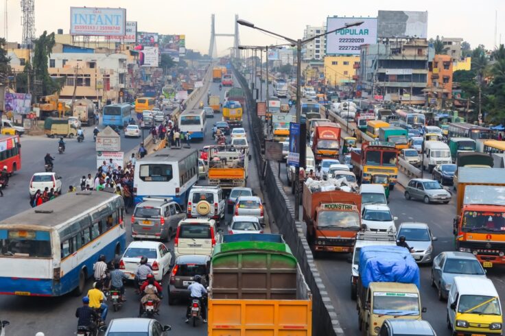 Congested urban highway at rush hour with cars, buses, and trucks crowded in multiple lanes while pedestrians stand on the roadside divider; billboards and buildings loom in the background.
