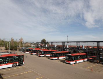 Multiple red and white city buses parked in a yard under a metal maintenance structure against a blue sky.