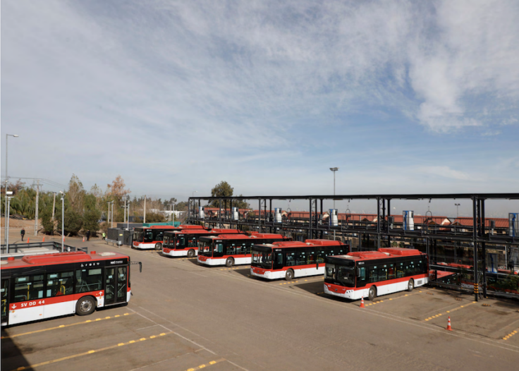 Multiple red and white city buses parked in a yard under a metal maintenance structure against a blue sky.