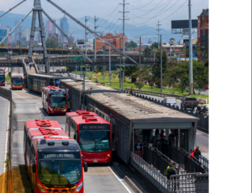 City transit hub with several red electric buses at a boarding platform and cable infrastructure against an urban skyline in the distance.