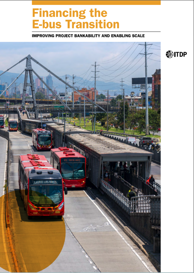 City transit hub with several red electric buses at a boarding platform and cable infrastructure against an urban skyline in the distance.