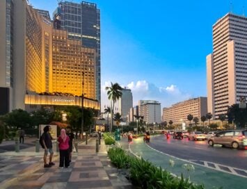 Urban street scene outside Grand Hyatt hotel with pedestrians, palm trees, and traffic at dusk/dawn.